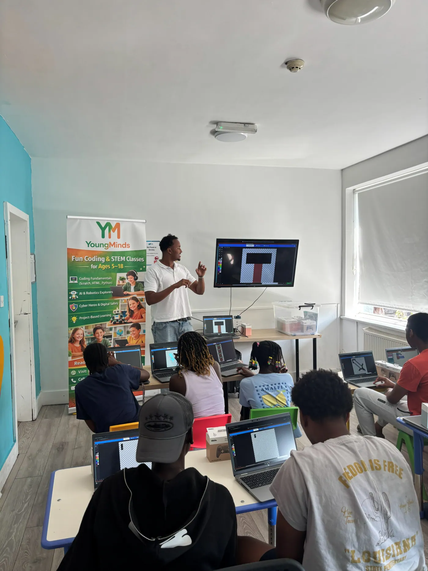 A mentor teaching sprite design to a coding club, with the YoungMinds banner and students at laptops